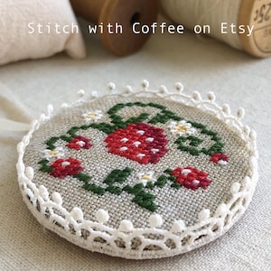 May include: A round, white, crocheted frame with a stitched design of red strawberries and green leaves on a beige linen background. The design is surrounded by a white crocheted border with small white beads.