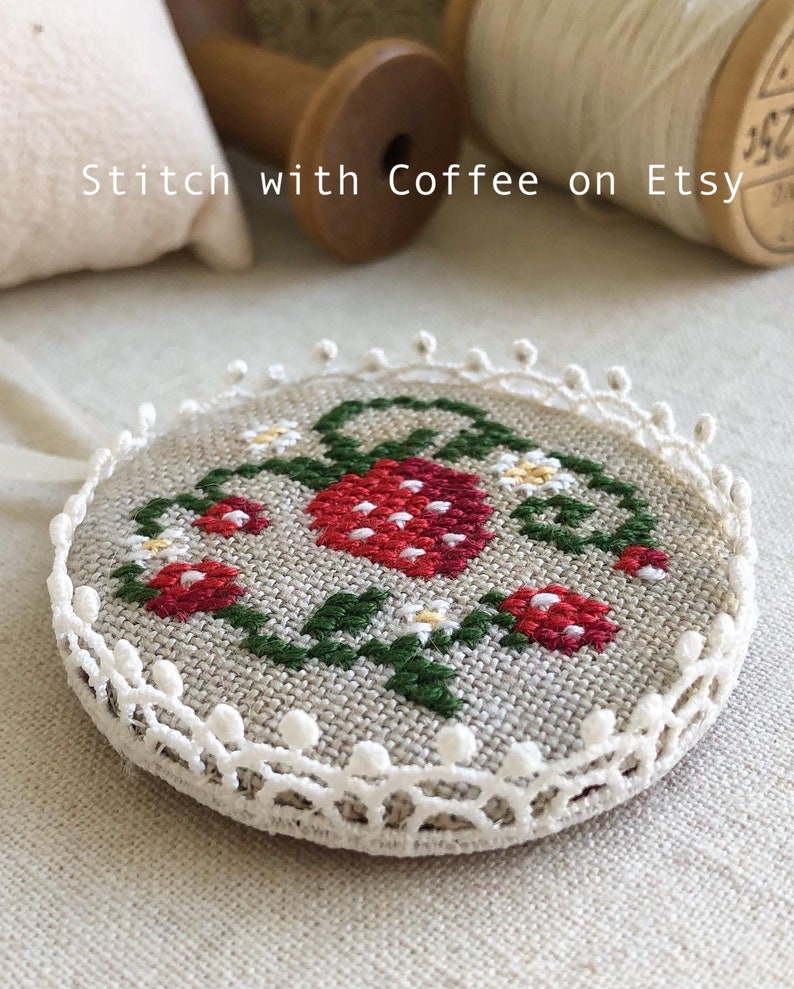 May include: A round, white, crocheted frame with a stitched design of red strawberries and green leaves on a beige linen background. The design is surrounded by a white crocheted border with small white beads.