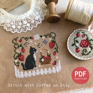 May include: Cross stitch pattern featuring a black cat sitting in a garden with strawberries, flowers, and a basket of strawberries. The pattern is on a beige linen fabric with a white picket fence at the bottom. The text "Stitch with Coffee on Etsy" is at the bottom of the image. The image also includes a spool of white thread, a wooden spool, and a piece of white lace.