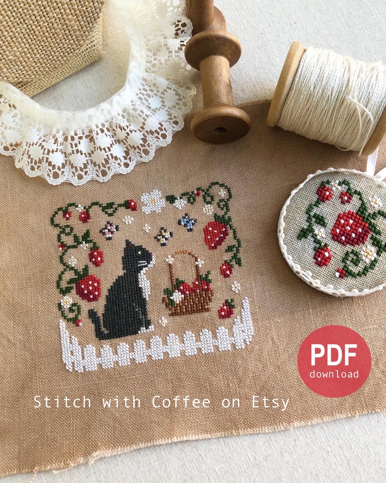 May include: Cross stitch pattern featuring a black cat sitting in a garden with strawberries, flowers, and a basket of strawberries. The pattern is on a beige linen fabric with a white picket fence at the bottom. The text "Stitch with Coffee on Etsy" is at the bottom of the image. The image also includes a spool of white thread, a wooden spool, and a piece of white lace.