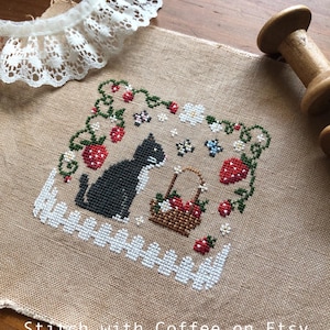 May include: Cross-stitch embroidery on a beige fabric with a black cat, a basket of strawberries, and a white picket fence. The design is surrounded by green vines and red strawberries. The fabric has the text "Stitch with Coffee on Etsy" at the bottom.