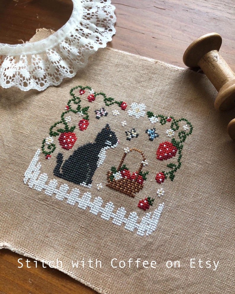 May include: Cross-stitch embroidery on a beige fabric with a black cat, a basket of strawberries, and a white picket fence. The design is surrounded by green vines and red strawberries. The fabric has the text "Stitch with Coffee on Etsy" at the bottom.