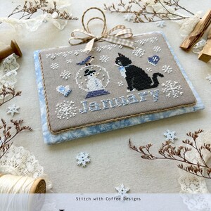 May include: A cross-stitch design featuring a black cat in a snow globe with a snowman inside. The design is on a beige fabric with blue trim and the word "January" is stitched in blue thread. The design is part of a monthly kitty calendar series for 2025.