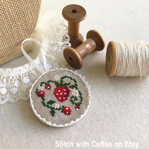 May include: A round, white-trimmed, linen ornament with a cross-stitch design of a red strawberry on a green vine with white flowers. The ornament is on a beige linen surface with two wooden spools and a spool of white thread.