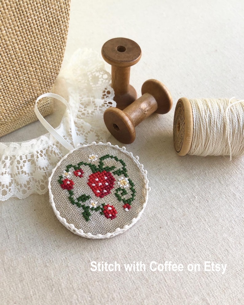 May include: A round, white-trimmed, linen ornament with a cross-stitch design of a red strawberry on a green vine with white flowers. The ornament is on a beige linen surface with two wooden spools and a spool of white thread.