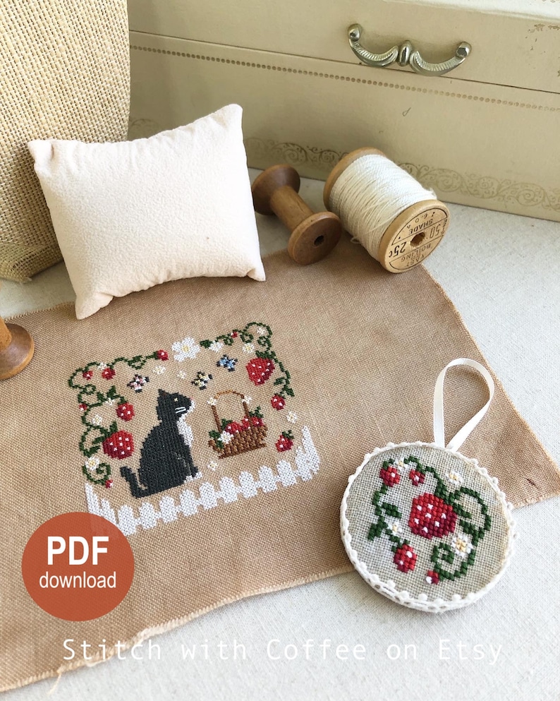 May include: Cross stitch pattern featuring a black cat sitting in a field of strawberries. The pattern is on a beige fabric and includes a small round ornament with a similar design. The text "PDF download" and "Stitch with Coffee on Etsy" are also visible.