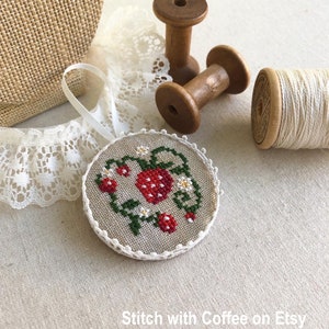 May include: A round, white, linen ornament with a cross-stitch design of a red strawberry with green leaves and white flowers. The ornament is hanging from a white ribbon and is surrounded by white lace and wooden spools of thread.