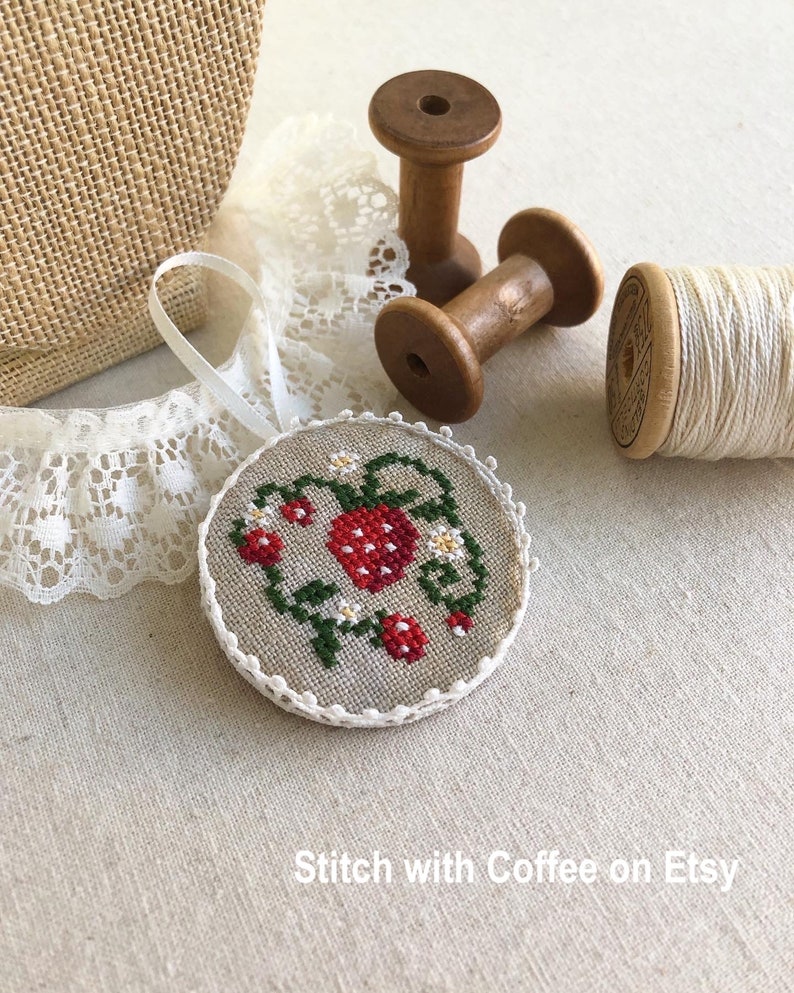 May include: A round, white, linen ornament with a cross-stitch design of a red strawberry with green leaves and white flowers. The ornament is hanging from a white ribbon and is surrounded by white lace and wooden spools of thread.