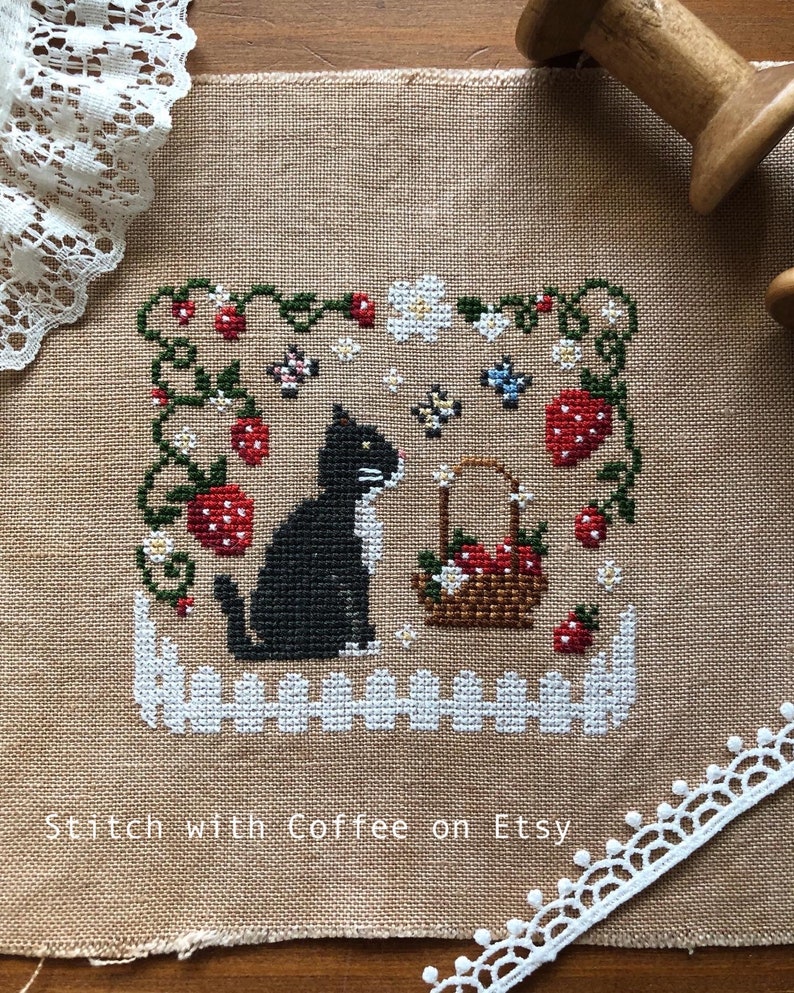 May include: Cross-stitch pattern of a black cat sitting in a garden with strawberries, flowers, and butterflies. The text "homestead" is stitched below the cat. The pattern is on a beige linen fabric.