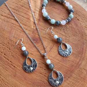 May include: A silver necklace, earrings, and bracelet set with gemstone beads. The necklace and earrings feature a teardrop-shaped pendant with a decorative pattern. The bracelet has a mix of green, gray, and pink beads. The jewelry is displayed on a wooden surface.