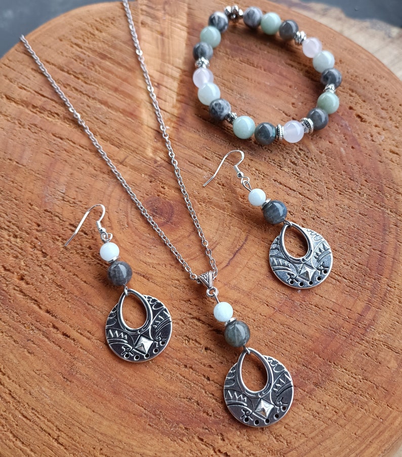 May include: A silver necklace, earrings, and bracelet set with gemstone beads. The necklace and earrings feature a teardrop-shaped pendant with a decorative pattern. The bracelet has a mix of green, gray, and pink beads. The jewelry is displayed on a wooden surface.