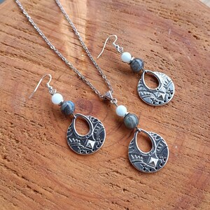 May include: A silver-toned jewelry set featuring a necklace and matching earrings. The set includes a pendant and two earrings with a teardrop-shaped design, adorned with intricate patterns and gemstone beads in shades of gray and white. The jewelry is displayed on a wooden surface.