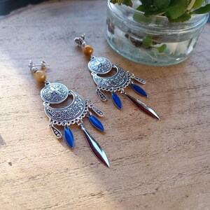 Op de afbeelding: Een paar zilverkleurige oorbellen met een bohemian design. Elke oorbel heeft een cirkelvormige bovenkant met een bloemmotief, een halvemaanvormig element, blauwe druppelvormige accenten en een lange, zilverkleurige hanger. De oorbellen hebben een lichtbruine kraal aan de bovenkant.