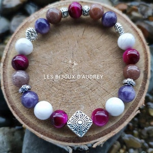 Puede incluir: Un brazalete hecho con cuentas de amatista, ojo de tigre y howlite blanca. El brazalete tiene un dije de plata con un diseño geométrico y las palabras "Les Bijoux d'Audrey".