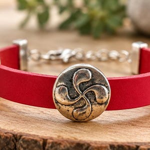 Könnte beinhalten: Ein rotes Lederarmband mit einem zentralen, kreisförmigen, silberfarbenen Anhänger mit einem vierarmigen, geschwungenen Design. Das Band wird durch zwei rechteckige, silberfarbene Metallendstücke mit Mustern und einen Kettenverschluss gehalten.