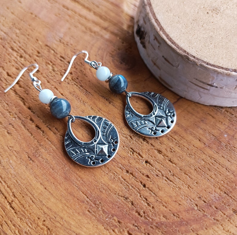 May include: Silver-toned dangle earrings with a teardrop-shaped pendant featuring an engraved design. Each earring has a white and a blue-grey bead. The earrings are displayed on a wooden surface.