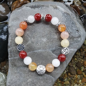 Peut inclure: Un bracelet composé de perles rouges, orange, blanches et jaunes avec des accents argentés. Le bracelet a un charme argenté avec un motif de nœud celtique.
