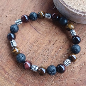 Puede incluir: Un brazalete de cuentas marrones, negras y doradas con detalles plateados. El brazalete está hecho de cuentas de roca volcánica, ojo de tigre y jaspe rojo.