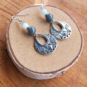 May include: Silver-toned dangle earrings with a teardrop-shaped pendant featuring an etched design. The earrings have gray and white beads. The earrings are displayed on a wooden surface.