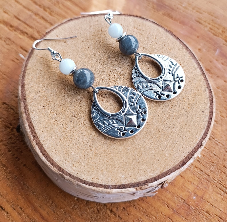 May include: Silver-toned dangle earrings with a teardrop-shaped pendant featuring an etched design. The earrings have gray and white beads. The earrings are displayed on a wooden surface.