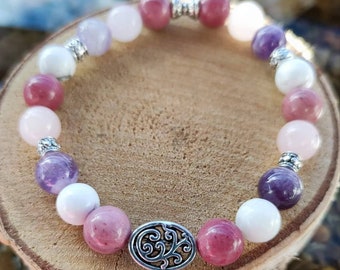 May include: A pink, purple, and white beaded bracelet with a silver charm featuring a floral design. The bracelet is made with natural stones and is designed to be worn on the wrist.