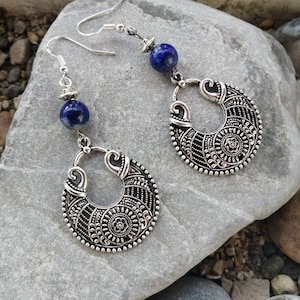 Peut inclure: Une paire de boucles d'oreilles créoles en argent avec une perle de lapis-lazuli bleue sur chaque boucle d'oreille. Les créoles ont un design détaillé et orné.