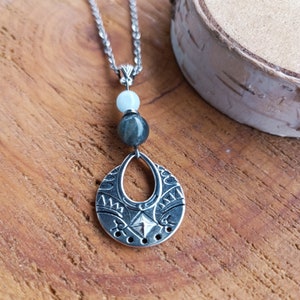 May include: Silver-toned pendant necklace with a detailed, openwork design. The pendant is paired with two gemstone beads and a delicate chain. The necklace is displayed on a wooden surface, showcasing its intricate details and craftsmanship.