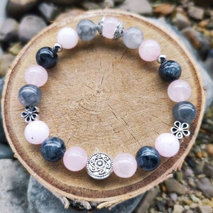 Könnte beinhalten: Ein Armband aus rosa, grauen und schwarzen Perlen mit silbernen Akzenten. Das Armband hat einen Blumenanhänger und einen runden Anhänger mit einem Design.