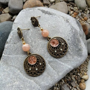 Peut inclure: Une paire de boucles d'oreilles pendantes avec une finition bronze. Chaque boucle d'oreille présente un pendentif rond et orné avec une pierre de couleur pêche au centre. Les pendentifs sont rehaussés de petites pierres blanches. Les boucles d'oreilles sont suspendues à une perle de couleur pêche et à un crochet bronze.