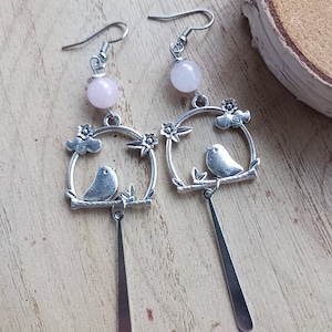 Op de afbeelding: Een paar zilveren oorhangers met een roze stenen kraal aan de bovenkant. De oorbellen hebben een vogel die op een tak zit met bloemen en wolken.