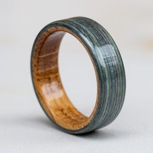 May include: A wooden ring with a unique design. The outer band is a muted teal color with a striped pattern, while the inner band is a light brown wood. The ring is a simple band style.