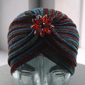 Op de afbeelding: Een veelkleurige turban met een glinsterende afwerking en een grote broche met een bloemversiering van edelstenen. De turban is op een glazen mannequinhoofd.