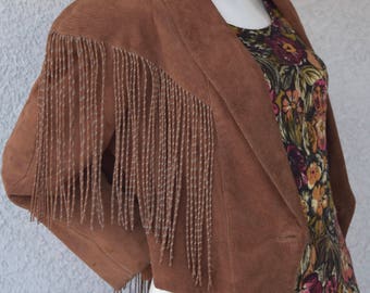 May include: A brown suede jacket with fringe detailing. The jacket has a long, flowing fringe that hangs down the sides and back. The jacket is worn over a floral dress.