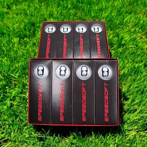 Puede incluir: Dos cajas abiertas de pelotas de golf sobre hierba verde. Cada caja contiene pelotas de golf con un diseño de bola ocho. Las cajas son negras con la palabra "SPEEDSOFT" en rojo. Las pelotas de golf son blancas.