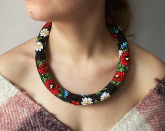 May include: A beaded necklace with a black background, adorned with red, white, and blue floral patterns. The necklace is a thick, circular design, showcasing a vibrant display of beadwork. The flowers are detailed with green leaves.