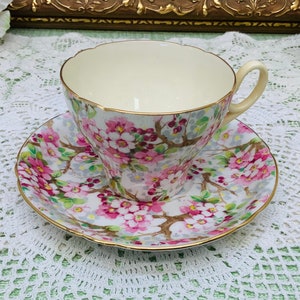 May include: A vintage teacup and saucer set featuring a floral design. The cup and saucer are decorated with pink and white flowers on a pale green background. The cup has a scalloped rim and a gold trim.