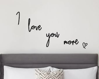 May include: Black metal wall decor with the words "I love you more" and a heart symbol. The decor is displayed above a gray headboard with white bedding and a patterned pillow.