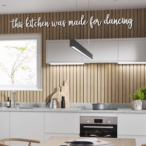 May include: A modern kitchen with white cabinets and a wood slat backsplash. The backsplash has the text "this kitchen was made for dancing" written on it. The kitchen has a stainless steel oven and a granite worktop.