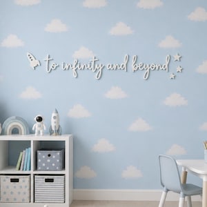 Peut inclure: Une enseigne murale blanche avec le texte en cursive to infinity and beyond, accompagn&eacute;e d'une petite fus&eacute;e et d'&eacute;toiles. L'enseigne est fix&eacute;e sur un mur bleu clair d&eacute;cor&eacute; d'un motif r&eacute;p&eacute;titif de nuages blancs.