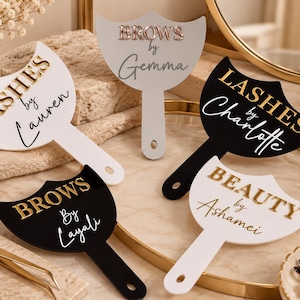 May include: Five handheld paddle mirrors in white and black with gold or rose gold 3D lettering. The mirrors feature custom text including LASHES by Lauren, BROWS by Gemma, LASHES by Charlotte, BROWS by Layali, and BEAUTY by Ashamei in various fonts.