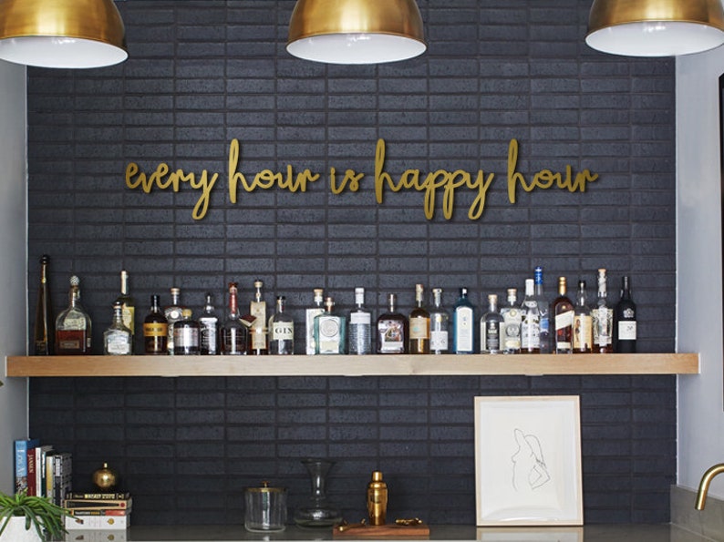 May include: A gold metal sign with the text "every hour is happy hour" mounted on a black brick wall above a shelf with a collection of liquor bottles.
