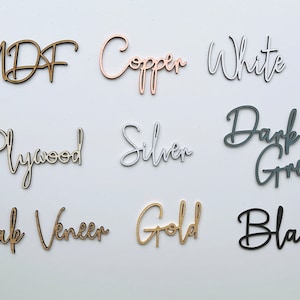May include: A collection of nine wood samples with their names written in cursive on each piece. The samples are in the following colors: MDF (natural wood), Copper (rose gold), White (white), Plywood (natural wood), Silver (silver), Dark Grey (grey), Oak Veneer (natural wood), Gold (gold), and Black (black).