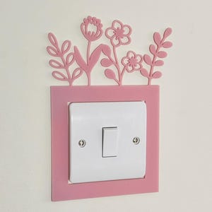 May include: A pink floral light switch cover with a white switch. The cover has a rectangular frame with decorative flowers and leaves at the top. The switch is centered within the frame, and the design is mounted on a white wall.