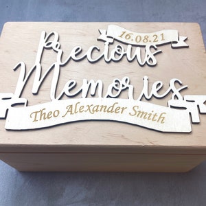 May include: A wooden box with a light wood finish. The lid has a cutout design that says "Precious Memories" with a banner that says "Theo Alexander Smith" and the date "16.08.21".