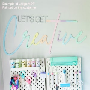 May include: A white pegboard with various craft supplies, including paint bottles, scissors, and tools. The pegboard is mounted on a wall with a pastel-coloured wooden sign that reads "LET'S GET CREATIVE".