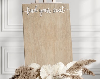 May include: A wooden sign with the text "find your seat" in white script. The sign is standing on an easel and is decorated with dried pampas grass and white paper fans.