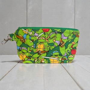 May include: Green zippered pouch featuring a repeating pattern of the Teenage Mutant Ninja Turtles. The pouch has a green zipper and a silver-colored clasp. The turtles are depicted in their signature colors: red, blue, purple, and orange.