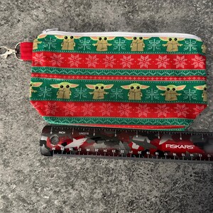 Puede incluir: Una bolsa rectangular con cremallera y un estampado festivo. El dise&ntilde;o presenta im&aacute;genes repetidas de un personaje de una popular serie de ciencia ficci&oacute;n, alternando con patrones de copos de nieve rojos y blancos sobre un fondo verde y rojo. Se adjunta un cierre plateado.