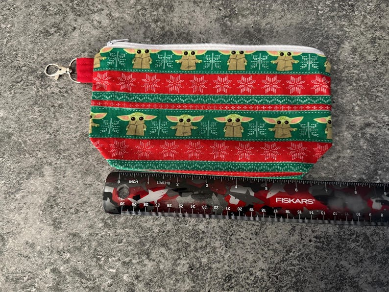 Puede incluir: Una bolsa rectangular con cremallera y un estampado festivo. El dise&ntilde;o presenta im&aacute;genes repetidas de un personaje de una popular serie de ciencia ficci&oacute;n, alternando con patrones de copos de nieve rojos y blancos sobre un fondo verde y rojo. Se adjunta un cierre plateado.