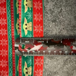Puede incluir: Una secci&oacute;n de tela con un patr&oacute;n festivo con tem&aacute;tica navide&ntilde;a. El dise&ntilde;o presenta rayas rojas y verdes alternas con motivos de copos de nieve y Grogu. Una regla muestra que la tela mide unos 5 cm de ancho.